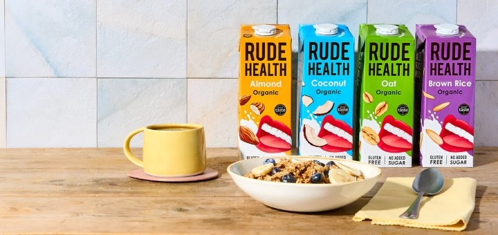 4 bottles of Rude Health milk alternatives on a bench next to some cereal and a coffee mug
