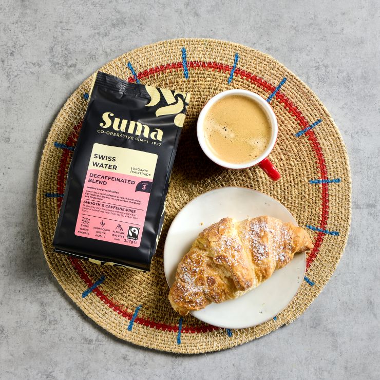 A birds eye view of a bag of Suma coffee, a cup of coffee and a croissant