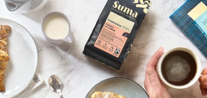 A bag of Suma Fair Trade Organic Honduran Ground Coffee on a table next to a cup of coffee and a pastry
