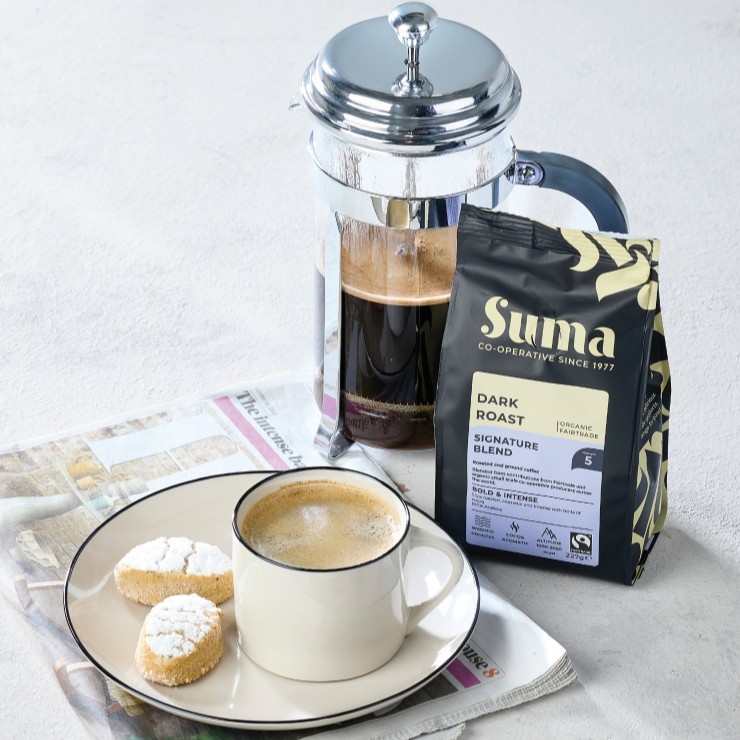 A bag of Suma Dark Roast coffee next to a filled french press and a cup of coffee