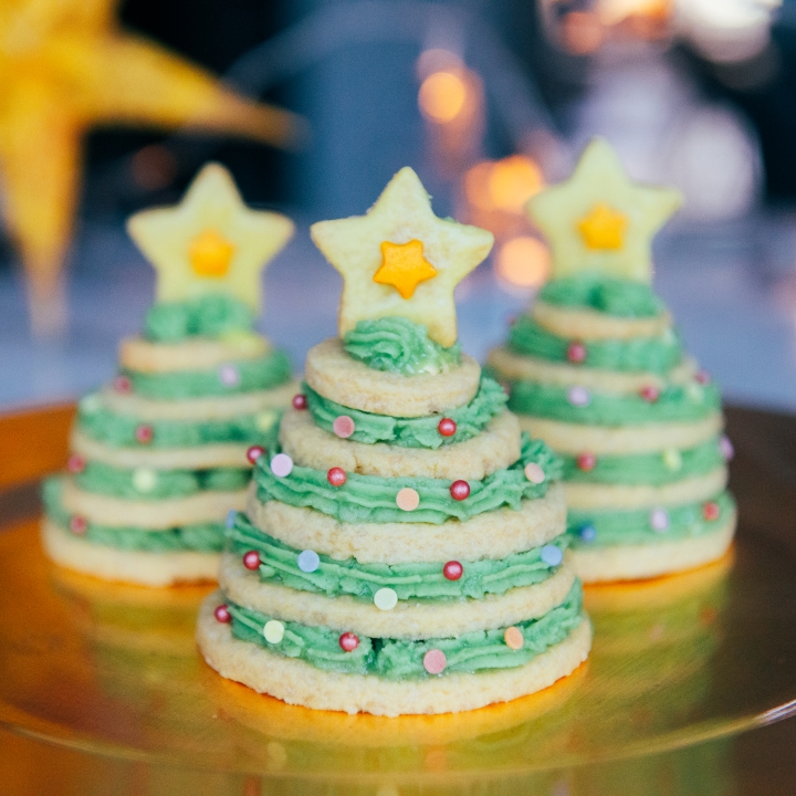 Three vegan Christmas tree biscuits on a table with blurred fairy lights in the background