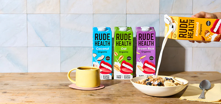 A hand pouring a carton of rude health almond milk into a bowl of cereal next to 3 other bottles of rude health milk alternatives.