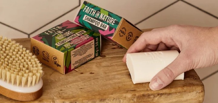 A hand holding a Faith in Nature shampoo bar next to a packaged shampoo bar and conditioner bar